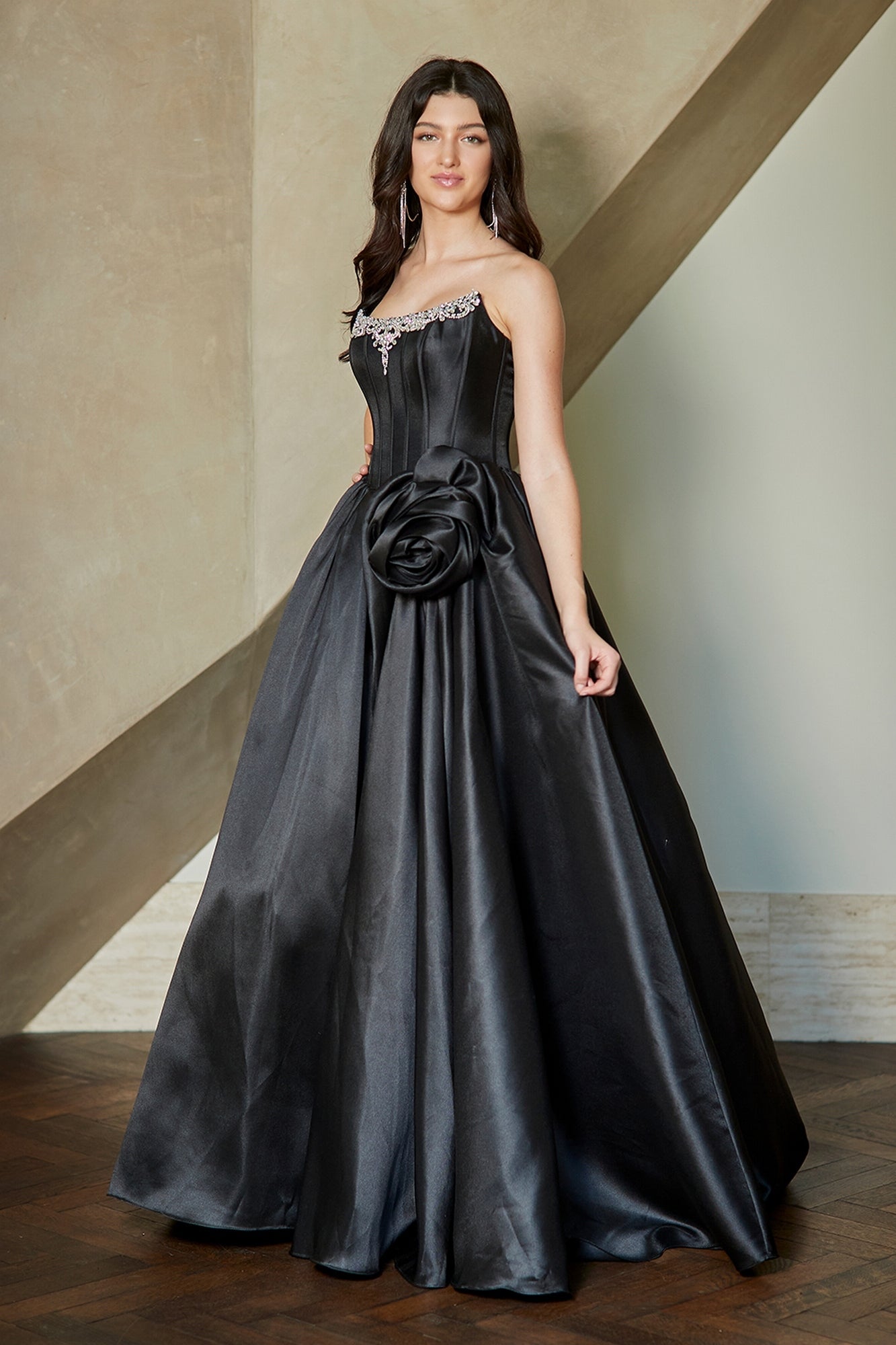 A person wearing a black Amelia Couture AL1401 corset prom ball gown with a rhinestone-embellished neckline and a large floral accent at the waist.